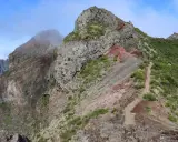 Steiler Bergpfad auf Madeira, ideal für die Packliste. Felsige Gipfel, grüne Vegetation und ein Weg, der sich durch die zerklüftete Landschaft schlängelt.