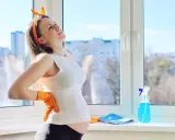 Ciężarna kobieta w pomarańczowych rękawiczkach z bólem pleców zastanawia się, czy w ciąży można myć okna. Obok butelka płynu i ściereczka.