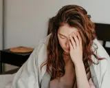 Kvinna med rött hår i sängen, täckt av filt, håller handen för ansiktet. Hon ser ut att ha feber, ett kolera symptom.