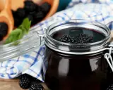Ein Glas mit dunklem Brombeergelee, garniert mit frischen Brombeeren. Daneben ein Holzschälchen mit weiteren Brombeeren und ein kariertes Tuch. Verschiedene Brombeergelee Varianten zum Genießen.