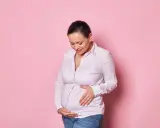 Kobieta w ciąży w wieku 45 lat z uśmiechem na twarzy delikatnie dotyka swojego brzucha.