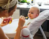 Mama karmi 7 miesięczne dziecko puree z marchewki. Maluch ma pomarańczowe ślady na buzi i śliniaczku.