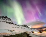 Abisko norrsken målar himlen i grönt och lila över snötäckta berg och en väg som leder mot upplysta hus.