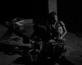 A woman in a patterned dress sits in shadow, eyes closed, while a man works with a bucket in the foreground. Alex Majoli captures a moment of quiet resilience.