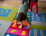 Dziecko wykonuje ćwiczenia na kolorowej macie z symbolami dłoni i stóp. Poduszka sensoryczna wspiera rozwój motoryczny.