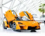 Ein leuchtend oranger McLaren Senna LM mit geöffneten Flügeltüren steht in einem modernen Gebäude mit Glasdach.