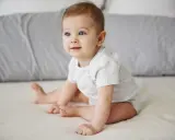 Niebieskooki niemowlak w jasnym body siedzi na łóżku, patrząc w bok.