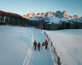 Quattro escursionisti camminano su un sentiero innevato al Passo di Lavazè, con le maestose vette dolomitiche illuminate dal sole al tramonto.