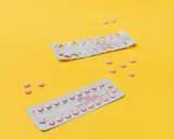Dos blísteres de pastillas anticonceptivas y varias pastillas sueltas, rosas, sobre fondo amarillo. Ayudan a regular la regla.