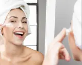 Donna sorridente con asciugamano in testa applica crema sul viso, parte della sua routine skincare.