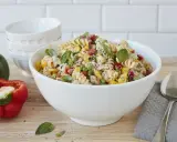 Ein bunter Nudelsalat ohne Mayonnaise mit Spiralnudeln, Erbsen, Mais, Paprika und frischem Basilikum in einer weißen Schüssel.
