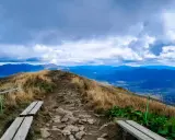 Kamienista ścieżka prowadzi przez trawiaste wzgórza, oferując zapierające dech w piersiach **Bieszczady widoki**. Na pierwszym planie drewniane ławki, w oddali błękitne pasma górskie pod burzowym niebem.