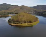 El meandro del Melero, una isla verde rodeada de agua serena, se abre paso entre colinas boscosas bajo un cielo azul.