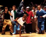 Energetyczna scena z filmów o tańcu. Breakdancer wykonuje akrobację, otoczony przez widzów i innych tancerzy.
