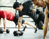 Drei Personen machen ein intensives Crossfit Training mit Kurzhanteln. Sie sind in einer Plank-Position und ziehen abwechselnd eine Hantel hoch.