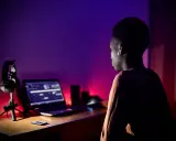 Un jeune homme, casque sur les oreilles, est assis devant un ordinateur portable, prêt à créer un son unique dans une ambiance tamisée aux lumières violettes et rouges.