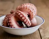 Pulpo listo para cocer en un cuenco blanco con borde azul, sobre una mesa de madera rústica.