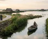 Ciclisti su un sentiero sterrato vicino a capanne di paglia e un pescatore in barca nel porto di Tolle.