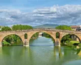 Der historische Puente la Reina überspannt mit seinen steinernen Bögen einen grünen Fluss. Dahinter erstreckt sich eine malerische Stadtkulisse.