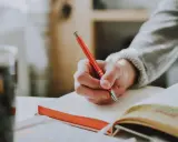 Main tenant une plume rouge, une main écrit dans un carnet. Un synonyme de participer pourrait être collaborer, comme dans cette scène d'étude.