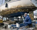 Un homme en combinaison bleue peint la partie immergée d'un bateau, recouverte de bernacles. Il utilise un rouleau pour appliquer une peinture bleue sur la coque.