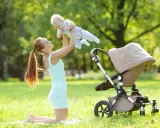 Uśmiechnięta mama podrzuca dziecko w powietrze na trawie obok wózka.