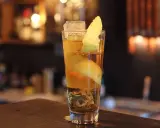 Ein Longdrink mit Eis, Zitronenzeste und einer Orangenscheibe. Die Zitrone windet sich wie ein Pferdehals im Glas.