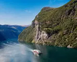 Ein Schiff fährt durch einen norwegischen Fjord im August. Das Wetter ist sonnig, die Berge sind grün und ein Wasserfall stürzt herab.
