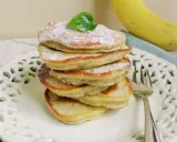 Stos puszystych, trzyskładnikowych placków bananowych, posypanych cukrem pudrem i udekorowanych listkiem, obok dojrzałego banana.