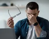 Homme fatigué, lunettes à la main, se frottant les yeux devant un ordinateur. Il est temps de penser à comment protéger ses yeux des écrans.