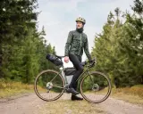 Wysoki mężczyzna w kasku i kurtce rowerowej pozuje na tle lasu, opierając się o swój gravelowy rower. Idealny rower dla wysokiego mężczyzny na leśne wyprawy.