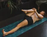 Femme effectuant un étirement psoas sur un tapis de yoga bleu. Elle est allongée sur le dos, une jambe pliée et ramenée vers la poitrine.