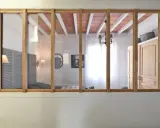 Une verrière en bois sépare deux pièces. On y voit un lit, des volets, une armoire et un lustre.