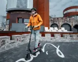 Un homme en casque et veste orange teste la vitesse de sa trottinette électrique devant un bâtiment de style médiéval moderne.