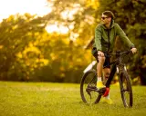Mężczyzna w zielonej kurtce i okularach przeciwsłonecznych odpoczywa na trawie obok swojego roweru elektrycznego ebike.