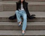 Donna con cappotto nero, camicia bianca e **pantaloni a sigaretta** chiari, indossa **tronchetti** blu.