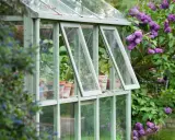 Une serre avec des fenêtres de récupération, remplie de plantes en pots, est entourée de lilas en fleurs.