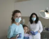 Uśmiechnięta studentka stomatologii w masce i rękawiczkach trzyma wiertło dentystyczne. W tle inne osoby w gabinecie.