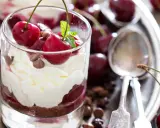 Delicioso postre con capas de crema batida, bizcocho de chocolate y cerezas frescas, coronado con más cerezas y trocitos de chocolate.