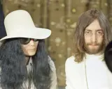John Lennon och Yoko Ono, ikoniska konstnärer, poserar tillsammans. Yoko bär en stor vit hatt och solglasögon, medan John har sitt karaktäristiska långa hår och glasögon.