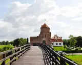 Drewniany most prowadzi do ceglanego zamku z czerwoną wieżą i białym budynkiem.