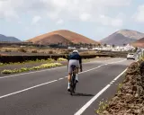 Ein Radfahrer auf einem Rennrad fährt auf Lanzarote durch eine vulkanische Landschaft mit weißen Häusern und Palmen.
