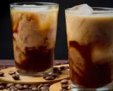 Två glas med iskaffe, perfekt för ett iskaffe recept. Krämig mjölk virvlar i mörkt kaffe med isbitar. Kaffebönor sprider sig runt glasen.