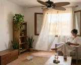 Mujer leyendo en sillón, con ventilador de techo y cortinas movidas por la brisa, una forma natural de como refrescar una habitación.