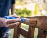 Mano aplicando barniz a un banco de jardín, creando un hermoso efecto madera.
