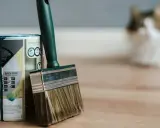 Puszka farby i pędzel czekają na malowanie paneli podłogowych. W tle rozmyty kot obserwuje przygotowania.