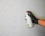 Mano con guante negro aplica spray para reparar gotelé. La pared tiene textura rugosa y blanca.