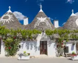 Trulli con simboli sui tetti, circondati da fiori e verde. Un'idea per scoprire **paesi da visitare vicino Alberobello**.