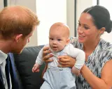 Meghan Markle håller sitt barn, som tittar nyfiket framåt. Prins Harry ser på dem med ett leende.