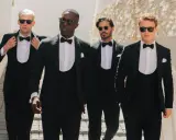Four men stride confidently in a modern black tie dress code, sporting sharp tuxedos and sunglasses.